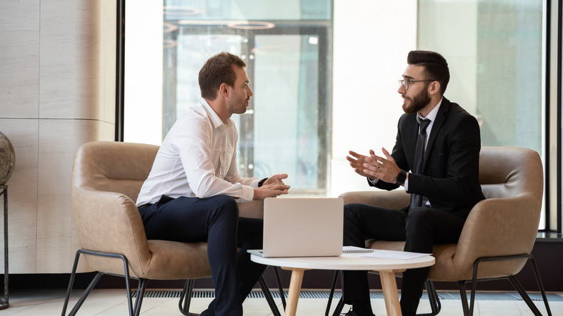 Middle eastern and caucasian ethnicity businessmen seated on armchair in modern office talking solve common issues, banker telling to client regarding bank services make recommendations and consulting